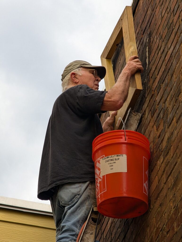 A volunteer on a ladder with a paint bucket, working on the brick exterior of the building.
