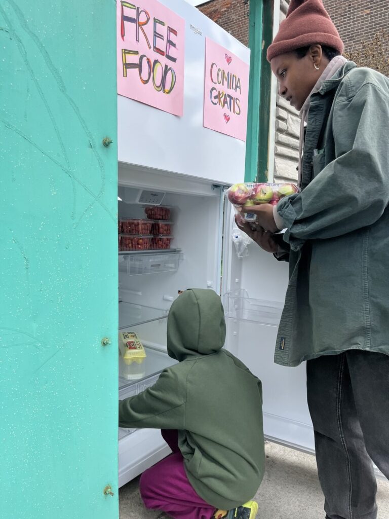 A neighbor stocking the Free Food Fridge outside TAUM while a child looks into the lower shelf.