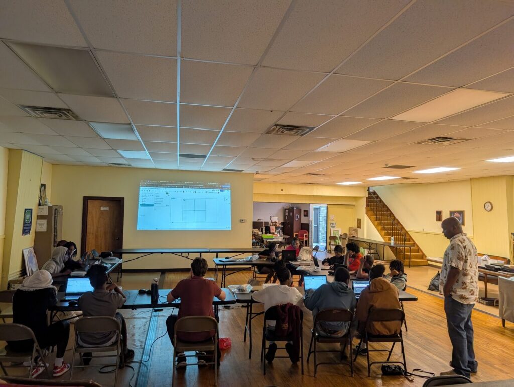 A class in progress in the community room, participants at laptops facing a projected screen.