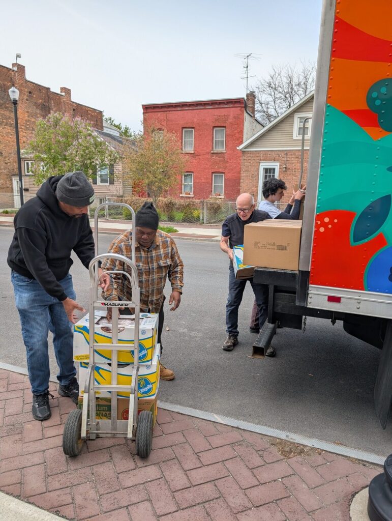 Volunteers unloading crates from a delivery truck onto a hand cart on the street.
